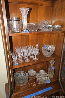 Photo showing multiple cut and pressed glass items including stemware, bowl, vase, and decanter inside a wooden cabinet.
