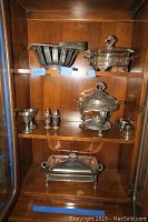 Photo showing multiple silver-plated serving pieces arranged in a wooden cabinet including chafing dishes, salt and pepper shakers, and small cups.