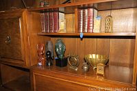 Wooden shelving with items: set of three carved head statues, several brass decorative pieces including bowl, apple, and Shalom decor, pair of stone bookends, brass style clock, and a set of books.