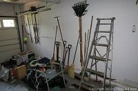 Wide view of yard tools and 6ft aluminum ladder leaning against the wall in garage showing arrangement and multiple tools such as rakes, shovels, brooms, and sport seat chair.