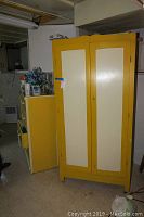 Photo of closed double door yellow and cream wooden armoire with a key inserted, showing basement surroundings.