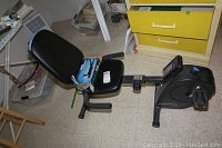 Recumbent bike with black padded seat and backrest, digital display, and pedals on tiled floor near yellow drawers and various home storage items.