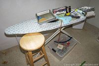 Photo of ironing board with baking pans, lint brush, wooden stool, sewing notion container, and boxed iron on top