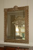 Full view of the large decorative mirror showing the ornate gold-toned carved frame and beveled glass reflecting a chandelier.
