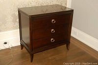 Front angled view of dark wood bedside cabinet with three drawers, brass ring pulls and tapered legs on hardwood floor
