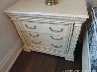 Front view of off-white three-drawer side table showing ornate carved moldings and silver-tone handles.