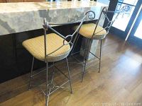 Photo showing two bar stools positioned by a kitchen counter with beige upholstered seats and decorative metal frames.
