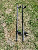 Two black metal roof rack crossbars laid out on grass, showing full length and attachment points.
