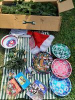 Overview of Christmas tree in box surrounded by holiday plates, garland, Santa hat, and small decorations on striped cloth outdoors.