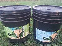 Two black plastic 5-gallon pails of Heavy Harvest garden fertilizer outdoors on grass, one labeled Spring Blend 22-11-11 and the other Summer Blend 17-17-17.
