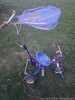 Photo of a blue child's walk behind tricycle with canopy, showing the full side view. The tricycle has three wheels, a molded plastic seat, handlebars with grips and a bell, pedals on the front wheel, and a blue fabric canopy over the seating area that shows signs of wear.