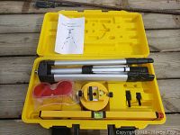 Open yellow carrying case showing laser level kit items including tripod, circular laser level, spirit level, red accessory box, two small attachments, and instruction manual.