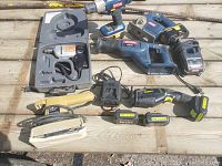 Photo showing a collection of power tools including Ryobi cordless drill and jigsaw, electric sander, corded drill in case, charger and two batteries on a wooden surface.