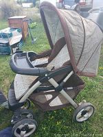 Brown and beige baby stroller shown outdoors on grass with canopy extended and plastic tray visible