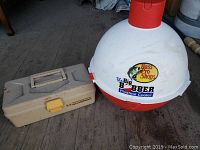 Photo showing a white and red spherical Bass Pro Shops Big Bobber Floating Cooler with a nearby brown Woodworm tackle box with yellow latch