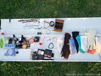 Top-down view of makeup items, nail polishes, jewelry, gloves and manicure accessories displayed on white panel outdoors on grass.