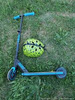 Youth two-wheeled scooter with blue grips and deck, alongside green and black spotted helmet on grass.