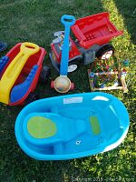 Outdoor photo of various baby and toddler toys on grass, including ride-on tractor, wagon, croquet mallet, bead maze, and baby bathtub