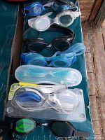 Seven pairs of adjustable swimming goggles displayed on a dark surface, including three new pairs in packaging.