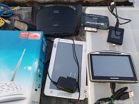 Photo showing Cisco Linksys wireless router, white E-reader with charger, TomTom GPS with charger, and a black device with cable.