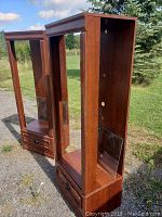 Two wooden display cabinets standing outdoors, showing front and side views. Each cabinet has glass shelves and bottom drawers.