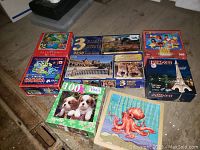 Seven assorted jigsaw puzzle boxes arranged on floor showing various themes, brands, and piece counts.