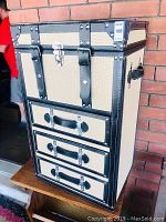 Front view of the dressing trunk showing three drawers with black leather-like handles and top compartment with latch lock, cream woven material with black trim and rivets.