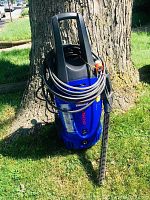 Blue Simoniz power washer standing upright against a tree with coiled hose and spray gun attached, seen from front-left angle.