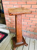 Full side view of the antique oak plant stand showing the square top and tapered column base with a wider base and three curved feet for stability, placed against a brick wall.