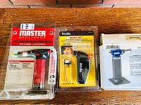 Three micro torches shown side by side on a wooden floor: Master Appliance MT-51 in red and black sealed package, Iroda Micro-Jet MJ-300 sealed package with lighter, and boxed Steinel torch.