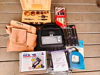 Full lot overview including leather tool belt, hole saw set in wooden box, screwdriver set, slot machine screwdriver box, Black & Decker case, tape measure, and accessories.