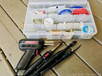 Overall view of soldering irons, soldering gun and plastic organizer box with spools of solder wire, electrical tape and rosin paste.