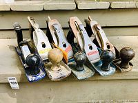 Full front view of six vintage hand planes arranged in a row showing various brands and knobs.
