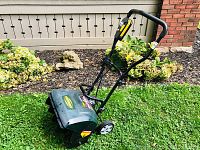 Side view of green electric snow thrower on grass near flowerbed featuring folding handle and wheels for maneuverability.