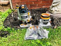 Wide view showing both routers and additional part in factory bag placed outdoors on grass and mulch next to a brick wall.