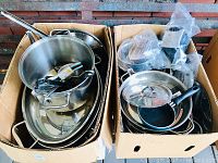 Top view of two cardboard boxes filled with stacked stainless steel pots, pans, lids, and kitchen utensils.