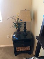 Photo of black side table with glass top, drawer open showing inside, wicker basket on bottom shelf, table lamp and artificial flowers placed on top