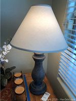 Table lamp with dark urn-shaped base and white conical shade on a wooden side table next to candles and a plant.