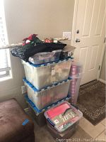 Stack of 4 large plastic totes with holiday decorations, wrapping paper rolls, and other items placed on the floor near the door.