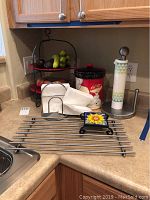 Photo of kitchen countertop showing a metal two-tier fruit stand with fruit (not included), a holiday-themed cookie jar with Santa Claus, a metal paper towel holder with paper towel, a metal drying rack, and a wire napkin holder with napkins.