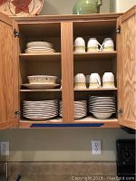 Photo of wooden cabinet with multiple shelves holding stacks of Studio Nova ceramic dishware including plates, bowls, and cups with floral designs.