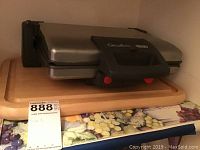 Electric grill placed on top of wooden cutting board with numbered lot tag in front.