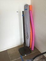 Umbrella stand, umbrella shade in gray fabric cover, and two colorful pool noodles positioned in corner against white wall and carpeted floor.