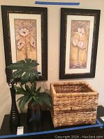 Photo showing two framed floral prints mounted on wall, faux plant in blue-tinted vase, two woven baskets stacked, and tall black lamp base without shade.