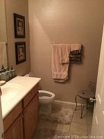 Photo showing a small decorative metal chair near the bathroom door, beige towels hanging on a wall rack, framed wall art on wall, beige bath rug in front of toilet and standard bathroom fixtures including a white toilet and sink with wooden cabinet.