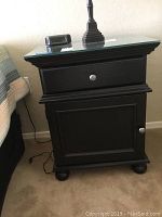 Front view of one black Thomasville nightstand showing glass top, drawer, cabinet door, bun feet, and silver knob.