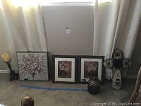 View of two matching framed flower prints, one framed canvas painting, a wooden candle holder with candle, woven pottery jar, and decorative wood and metal cross laid out on floor.