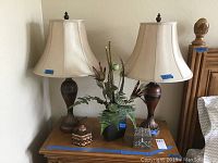 Photo showing two wooden lamps with beige lampshades, silk flower arrangement in black vase, and two small trinket boxes on wooden surface.