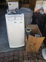 Front and angled view of white EnStar portable air conditioner next to a cardboard box containing vent hose and accessories.