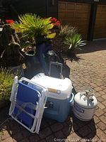 Lot shown outdoors on paved area with plants and garage door in background, displaying cooler, propane tank, and folded chairs.